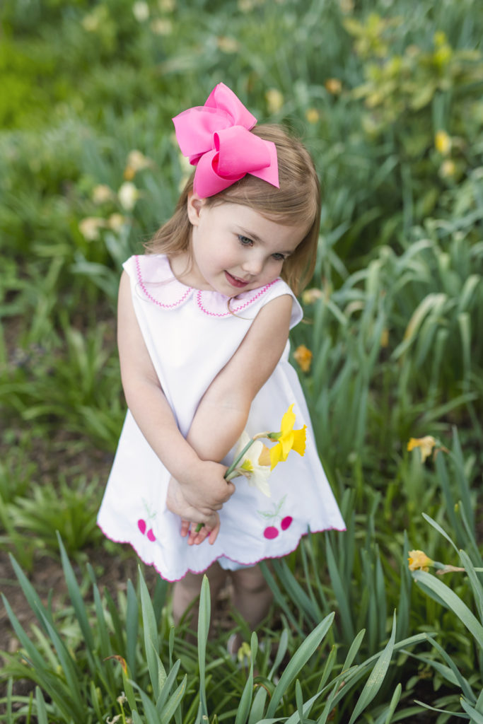 Morven Park in Loudoun County. Toddler girl wearing a cherry romper from the perfect peony, purchased at Ashby Mae Boutique. She holds a yellow daffodil 