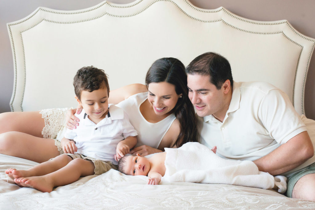 Caucasian family of mom, dad and big brother dressed all in white dote over their newborn baby girl while lying on a modern white bed.
