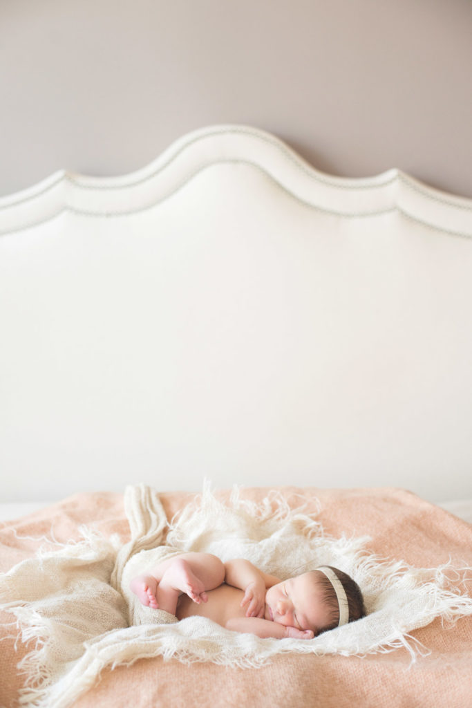 newborn baby girl lying on bed on a peach blanket and wearing white headband