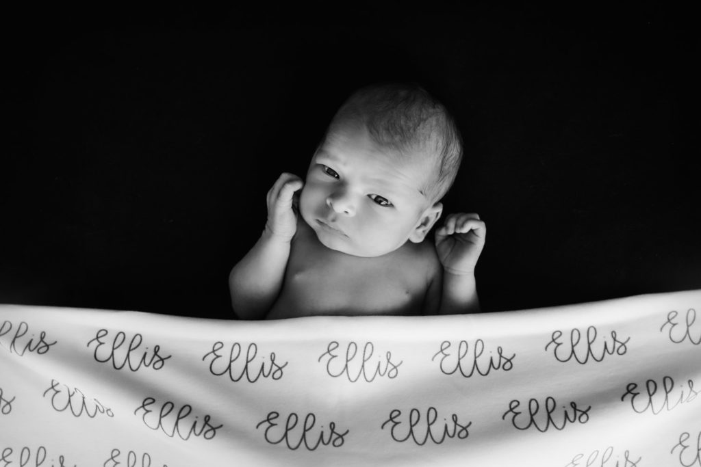 black and white image of Caucasian newborn baby boys with eyes wide open lying under a personalized name blanket