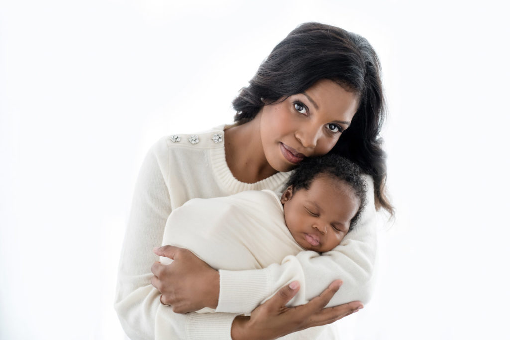 Black mom with dark hair and eyes wearing a cream sweater and holding her newborn baby 
swaddle in a cream wrap