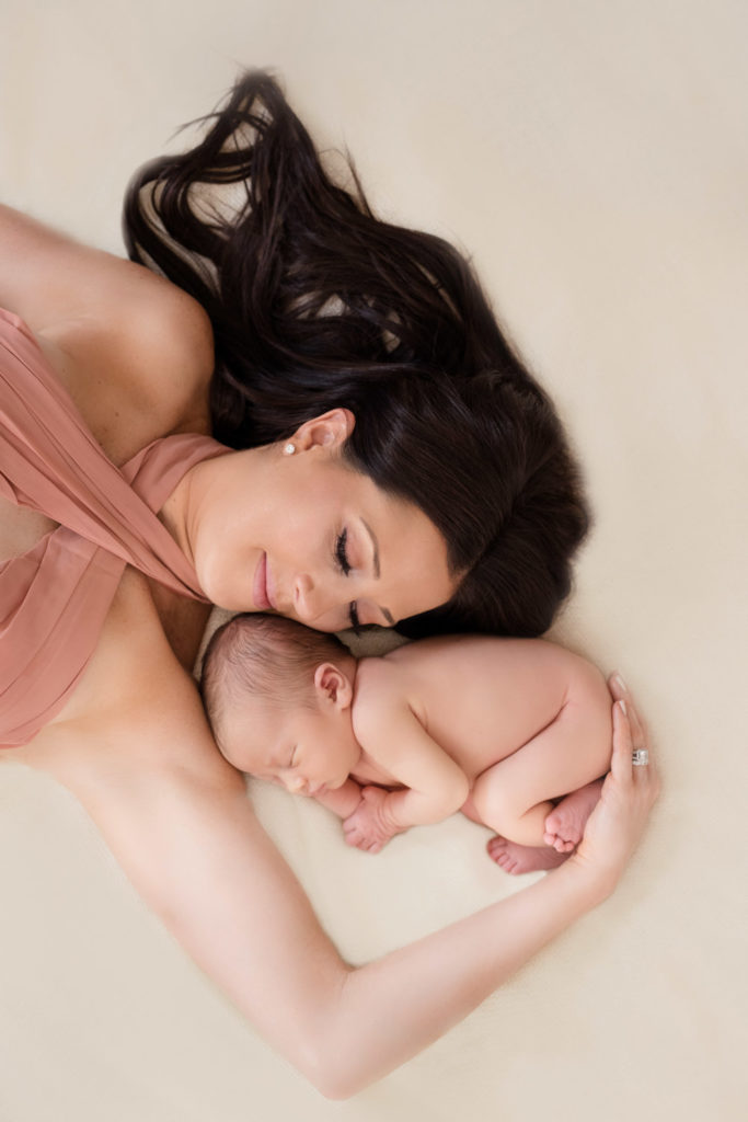 Caucasian mom wearing a blush halter chiffon dress lies on a cream background holding her newborn baby girl 