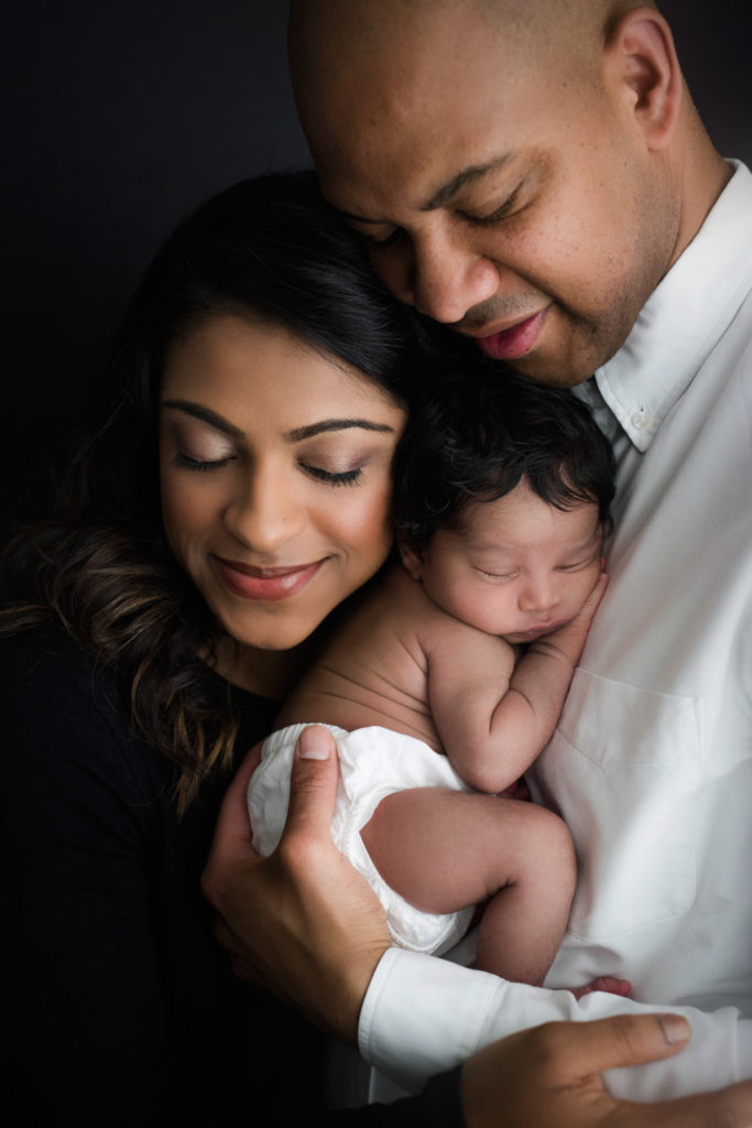 Close up image from newborn photography session of black family with dad dressed in white button down top and mom dressed in all black holding their baby boy