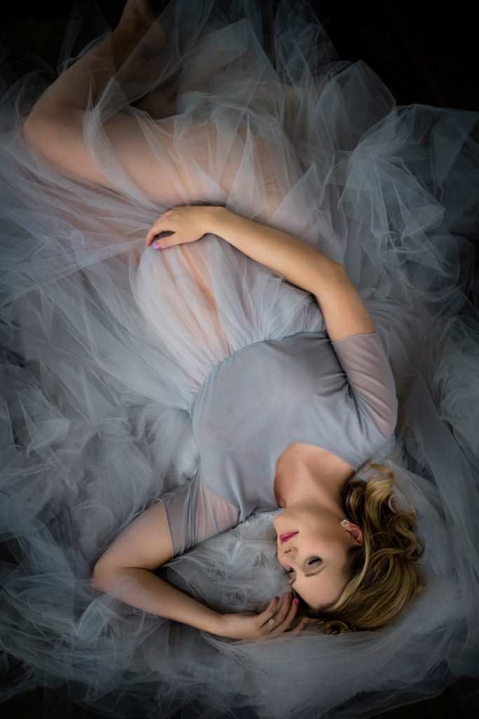 A glamour maternity photo with mom surrounded in pale blue tulle laying on the ground with hand on her belly