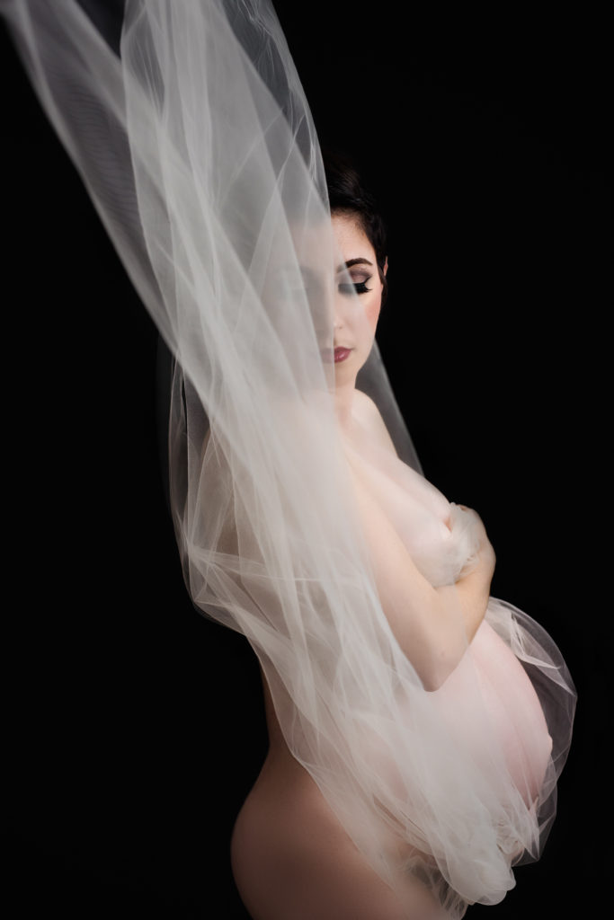 Expectant mother draped in white tulle in front of all black background in dramatic lighting 