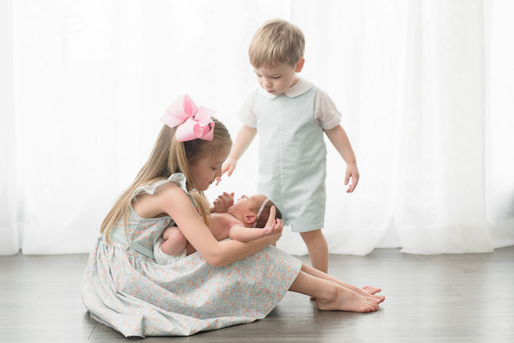 Young girl holds her newborn sister while their toddler sibling looks down at them for photos