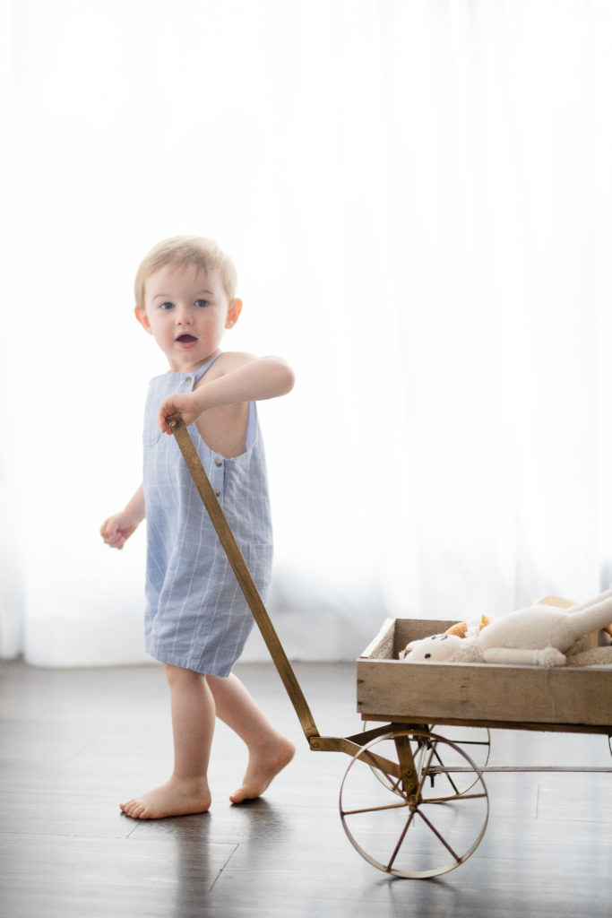 Toddler sibling plays with wooden cart in a photo studio during newborn photos with a white curtain behind him