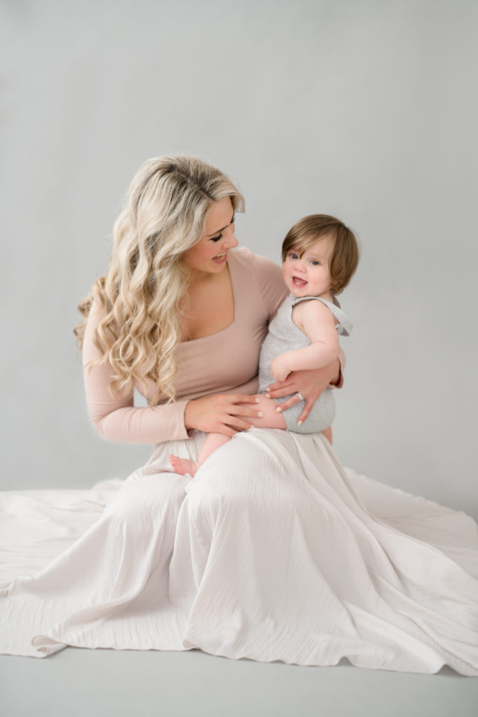 A blonde mom with long hair poses with her baby in a gray romper in a photo studio in Leesburg