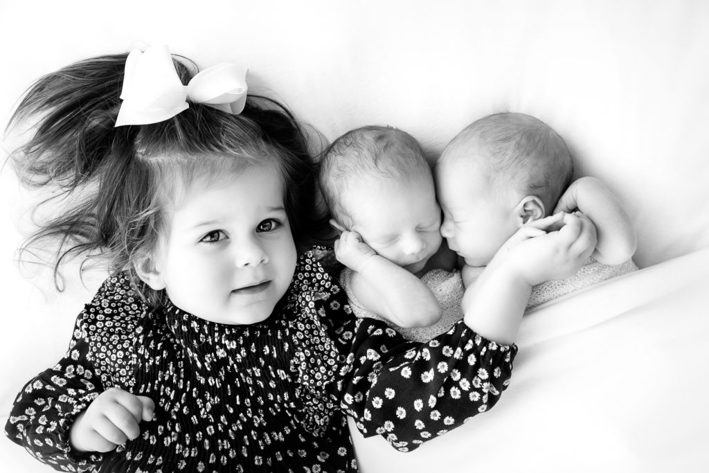 [Black and white photo of a toddler older sibling snuggling with her newborn twin siblings]