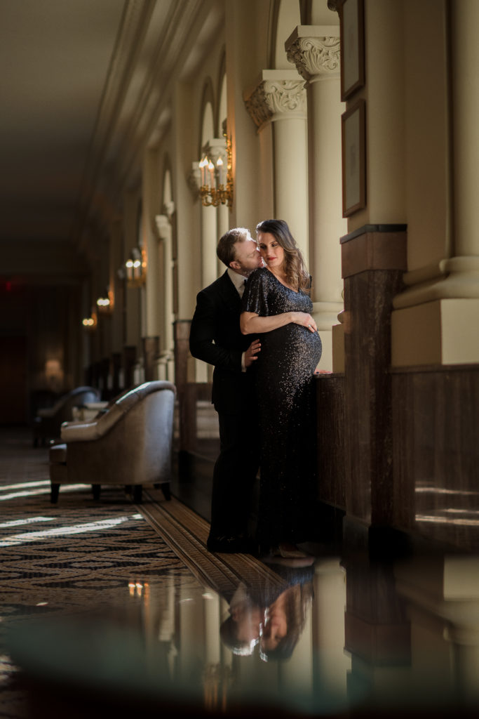 man stands behind wife and kisses her cheek at hotel in DC for maternity photo session