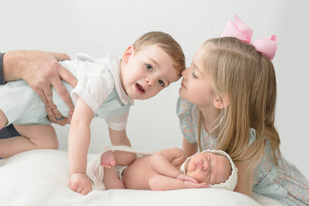 portrait by Leesburg Photographer of toddler boy leans into frame while big sister cradles newborn baby and kisses toddler for newborn milestone 