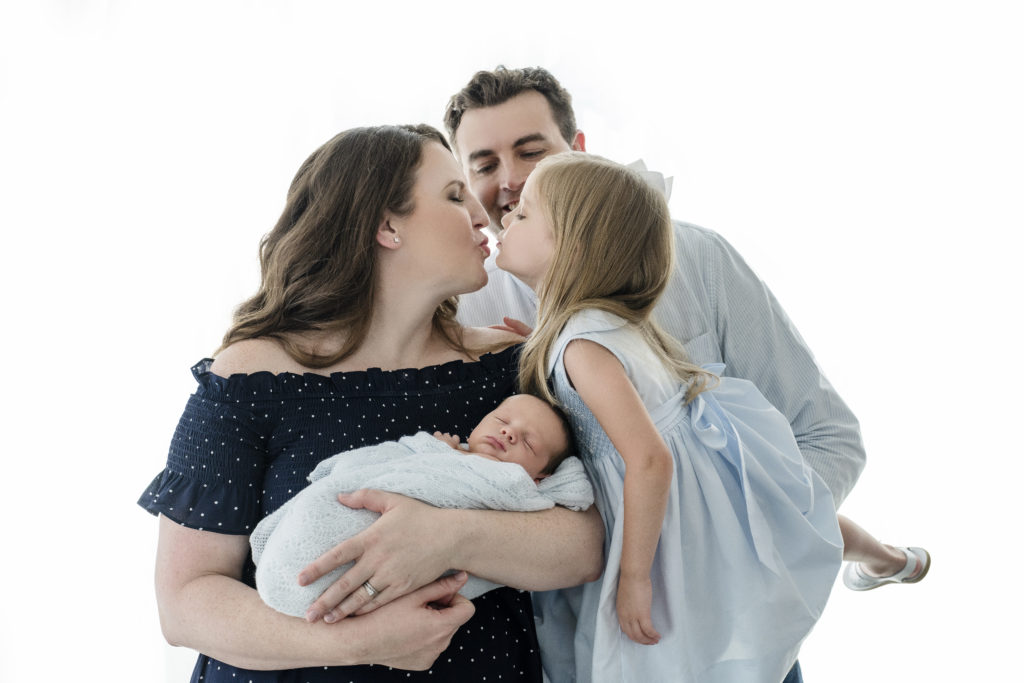 Mom holds newborn and dad holds toddler kissing mom