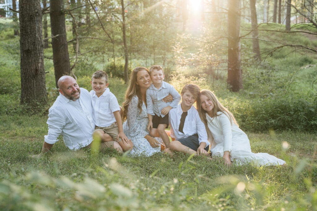 Family of six sits in tall grass with sun streaming in for family photos